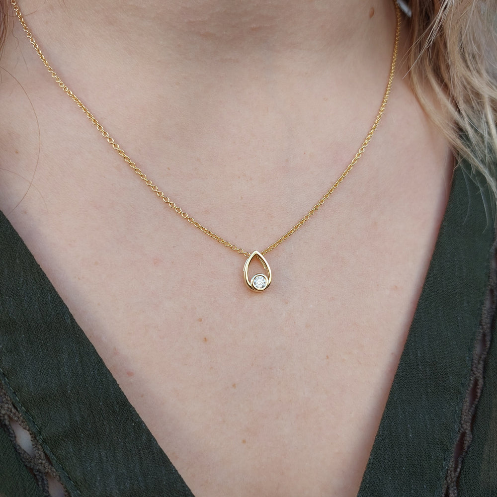 A product photo of a moissanite pendant in yellow gold sits on a clear white background. The pendant is in the shape of a small teardrop, with a petite gold-encased circle-cut moissanite gem nestled at its base. The yellow gold teardrop is smooth and unblemished. 