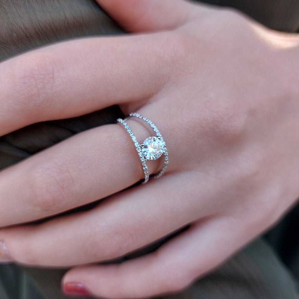 
            
                Load image into Gallery viewer, A product picture of a brilliant white moissanite engagement ring sitting on a woman&amp;#39;s finger. The central moissanite jewel reflects several vibrant colours off of its many angles, while white jewelled detailing hugs the rims of the split 9k white-gold band.
            
        