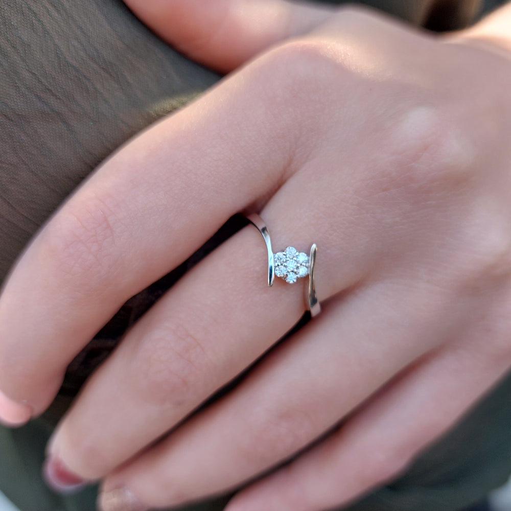 A product photo of a moissanite ring in 9k white gold on a woman's finger A simple crescent band hugs the top and bottom of the 7-jewel cluster of reflective moissanite gems in the centre.