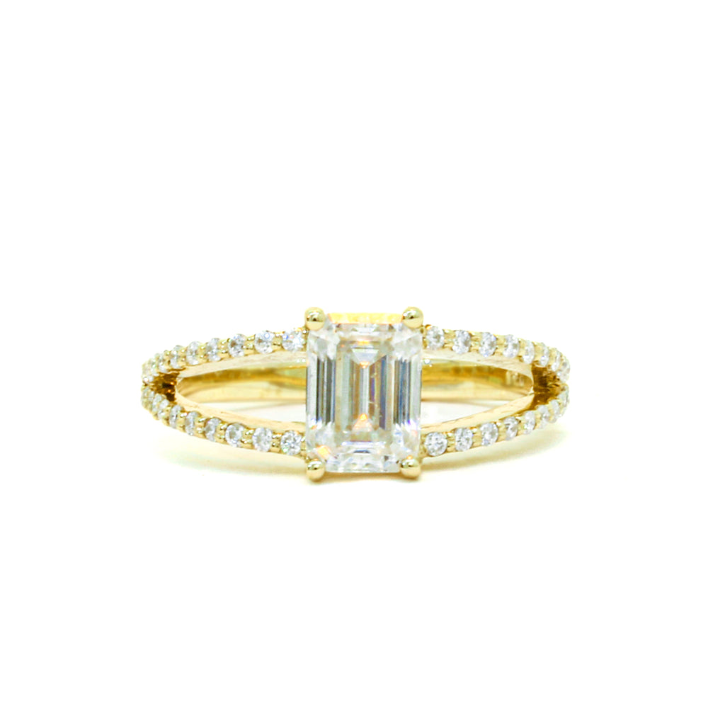 A product photo of a moissanite ring in 9k yellow gold sitting on a clear white background. The reflective emerald-cut moissanite jewel sits in the centre. A simple yellow gold band becomes embedded with diamonds the closer it gets towards the jewel, and splits into two bands on either side, both meeting in the centre of the ring to hold the moissanite in place.