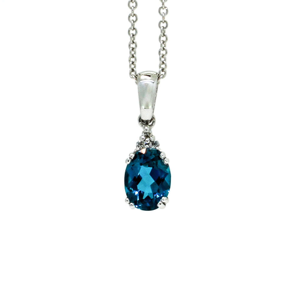 A product photo of a white gold london blue topaz pendant sitting against a white background. The deep blue oval-cut stone is held in place by 4 pairs of claws at its top and bottom, with a trio of white diamonds connecting the stone to the rest of the pendant. It is suspended by a simple white gold chain.
