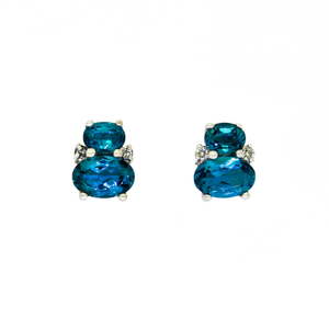 A product photo of a pair of white gold london blue topaz earrings sitting against a white background. One smaller oval is stacked upon a larger oval on each earring, with the area where the two stones make contact decorated by a single small diamond on either side.