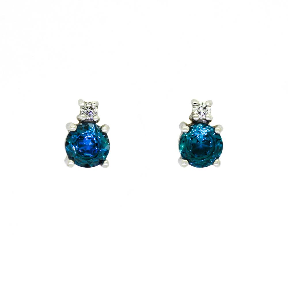 A product photo of a pair of white gold london blue topaz earrings sitting against a white background. The simple circle-cut stones are complimented by a single white diamond atop each stone, encased in white gold.