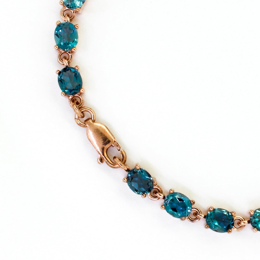 A product photo of a rose gold bracelet made up of many oval-shaped London Blue Topaz gems sitting on a clear white background.