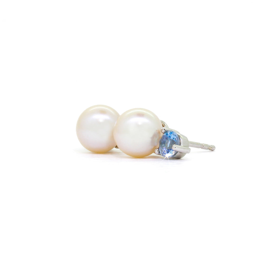 A product photo of delicate white pearl earring studs on a white background. The rosaline pearls each have aquamarine detailing above them connecting to the white gold studs.