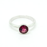 A product photo of a silver ring with a bezel-set rhodalite centre stone sitting on a white background. The silver band is simple and smooth, connecting on either side of a circle-cut rhodalite stone surrounded by a solid frame of silver. The rhodalite jewel is a deep velvet purple colour, reflecting a warm plum colours across its multi-faceted surface