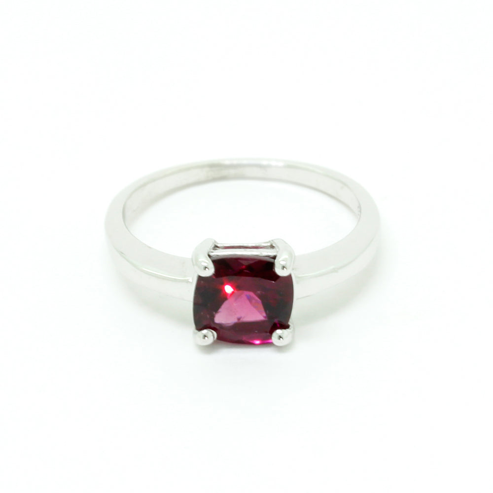 A product photo of a silver ring with a cushion-cut rhodalite centre stone sitting on a white background. The silver band is simple and smooth, connecting on either side of a square cushion-cut rhodalite stone held in place by four delicate silver claws. The rhodalite jewel is a deep velvet purple colour, reflecting a warm plum colours across its multi-faceted surface.
