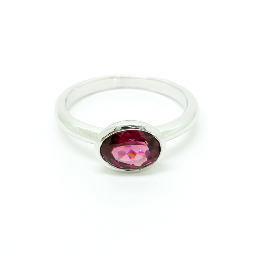 A product photo of a silver ring with a bezel-set rhodalite centre stone sitting on a white background. The silver band is simple and smooth, connecting on either side of a horizontally-oriented oval-cut rhodalite stone surrounded by a solid frame of silver. The rhodalite jewel is a deep velvet purple colour, reflecting a warm plum colours across its multi-faceted surface.