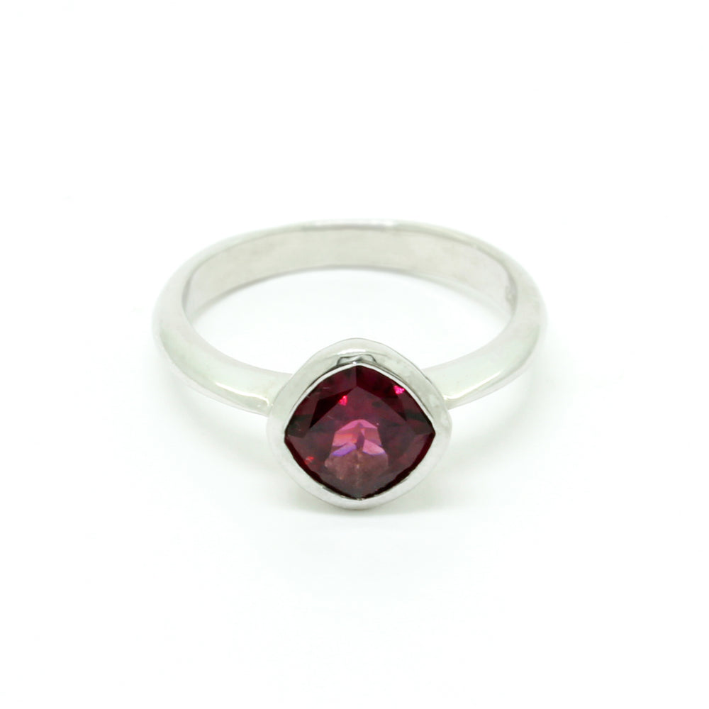 
            
                Load image into Gallery viewer, A product photo of a silver ring with a bezel-set rhodalite centre stone sitting on a white background. The silver band is simple and smooth, connecting on either side of a diagonally-oriented cushion-cut rhodalite stone surrounded by a solid frame of silver. The rhodalite jewel is a deep velvet purple colour, reflecting a warm plum colours across its multi-faceted surface.
            
        
