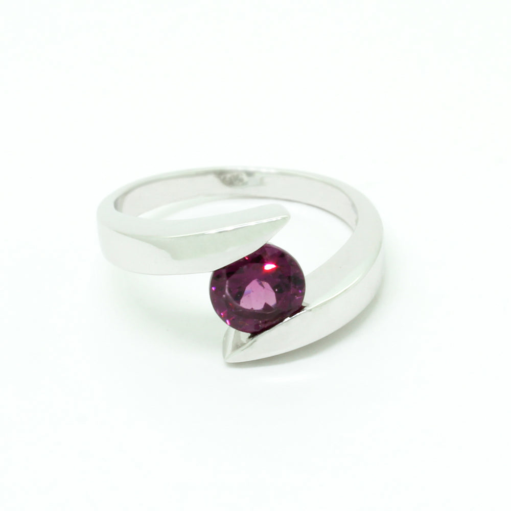 A product photo of a silver ring with a circle-cut rhodalite centre stone sitting on a white background. The reflective round-cut rhodalite sits in the centre, and a smooth silver band splits on either side of the jewel, hugging the top and bottom of the stone to hold it in place. The rhodalite jewel is a deep velvet purple colour, reflecting a warm plum colours across its multi-faceted surface.