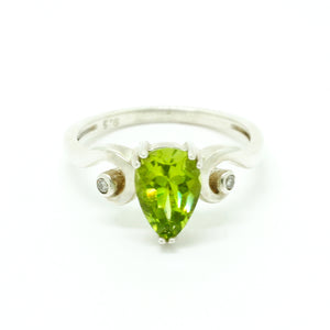 A product photo of a silver ring with a pear-cut peridot centre stone sitting on a white background. The silver band is simple and smooth before coiling elegantly on either side of the peridot centre stone. The centre of each silver coil is set with a tiny diamond. The peridot jewel is a shade of bright, vibrant green, reflecting chartreuse light across its multi-faceted surfaces.