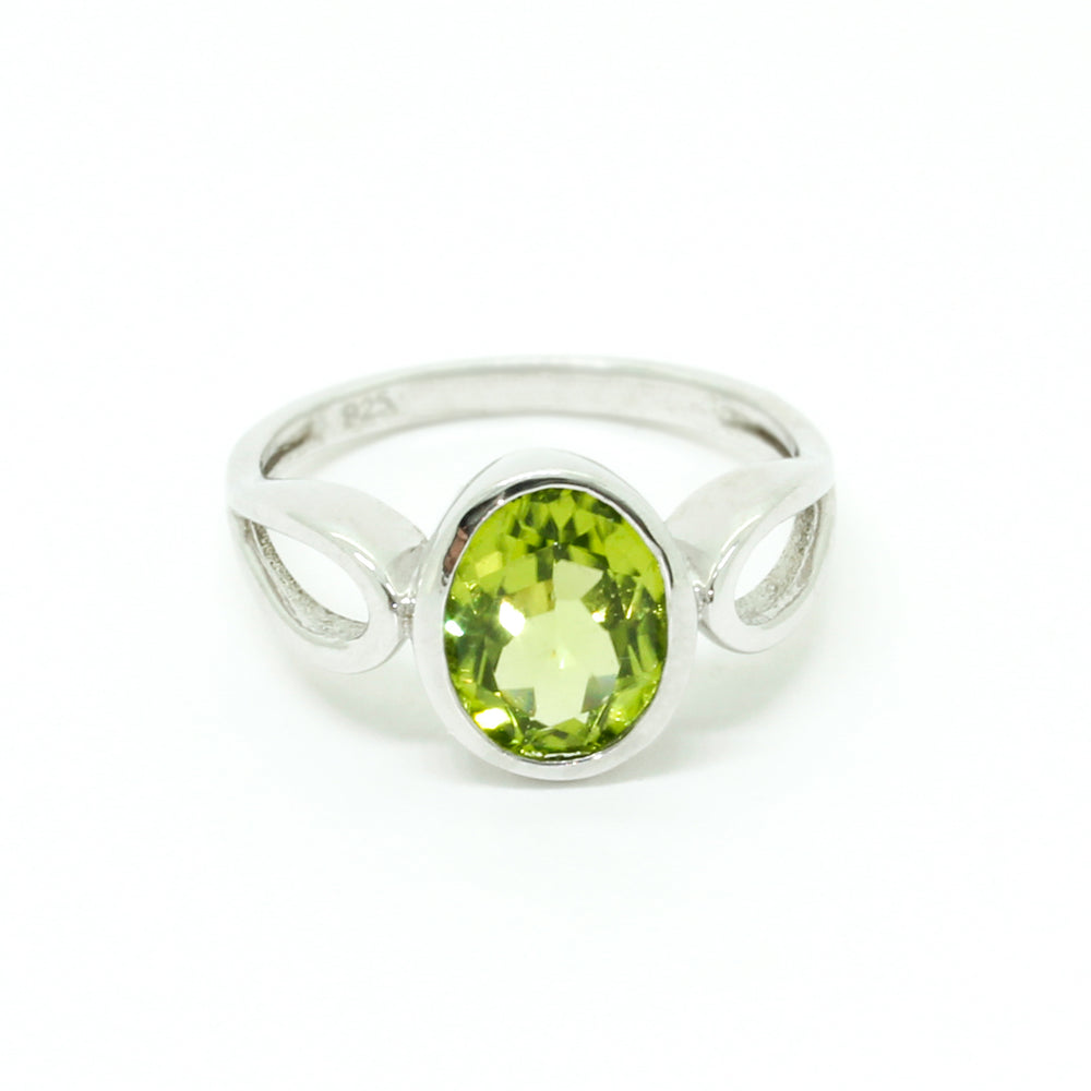 A product photo of a silver ring with an bezel-set oval-cut peridot centre stone sitting on a white background. The silver band is simple and smooth before splitting into delicate loops on either side of the peridot centre stone. The peridot jewel surrounded by a solid frame of silver and is a shade of bright, vibrant green, reflecting chartreuse light across its multi-faceted surfaces.