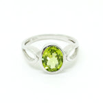 A product photo of a silver ring with an bezel-set oval-cut peridot centre stone sitting on a white background. The silver band is simple and smooth before splitting into delicate loops on either side of the peridot centre stone. The peridot jewel surrounded by a solid frame of silver and is a shade of bright, vibrant green, reflecting chartreuse light across its multi-faceted surfaces.