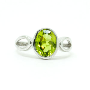 
            
                Load image into Gallery viewer, A product photo of a silver ring with an bezel-set oval-cut peridot centre stone sitting on a white background. The silver band is simple and smooth before splitting into delicate loops on either side of the peridot centre stone. The peridot jewel surrounded by a solid frame of silver and is a shade of bright, vibrant green, reflecting chartreuse light across its multi-faceted surfaces.
            
        