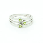 A product photo of a silver ring with 3 delicate bezel-set peridot stones sitting on a white background. The silver band is simple and smooth at the back before splitting into three prongs, each capped off with a delicate little peridot stone set within a silver frame. The prongs end at three different lengths, creating an interesting connecting line between the three stones. The peridot jewels are a shade of bright, pale green.