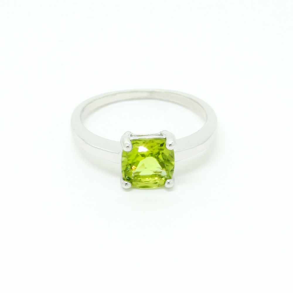 A product photo of a silver ring with a cushion-cut peridot centre stone sitting on a white background. The silver band is simple and smooth, connecting on either side of a square cushion-cut peridot stone held in place by four delicate silver claws. The peridot jewel is a shade of bright, vibrant green, reflecting chartreuse light across its multi-faceted surfaces.