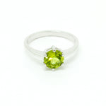 A product photo of a silver ring with a round-cut peridot centre stone sitting on a white background. The silver band is simple and smooth, connecting on either side of the circular peridot stone held in place by six silver claws. The peridot jewel is a shade of bright, vibrant green, reflecting chartreuse light across its multi-faceted surfaces.