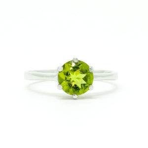 A product photo of a silver ring with a round-cut peridot centre stone sitting on a white background. The silver band is simple and smooth, connecting on either side of the circular peridot stone held in place by six silver claws. The peridot jewel is a shade of bright, vibrant green, reflecting chartreuse light across its multi-faceted surfaces.