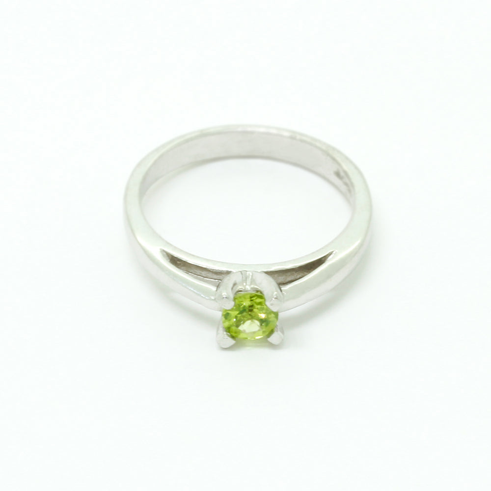 A product photo of a silver ring with a round-cut peridot centre stone sitting on a white background. The silver band is simple and smooth, connecting on either side of a small, roundl-cut peridot stone, held in place by four silver claws and a split band behind it. The peridot jewel is a shade of bright, vibrant green, reflecting chartreuse light across its multi-faceted surfaces.