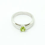 A product photo of a silver ring with a round-cut peridot centre stone sitting on a white background. The silver band is simple and smooth, connecting on either side of a small, roundl-cut peridot stone, held in place by four silver claws and a split band behind it. The peridot jewel is a shade of bright, vibrant green, reflecting chartreuse light across its multi-faceted surfaces.