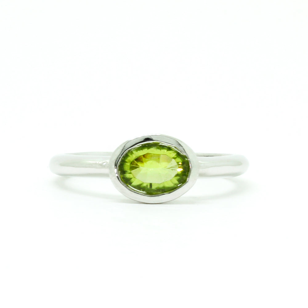 A product photo of a silver ring with a bezel-set peridot centre stone sitting on a white background. The silver band is simple and smooth, connecting on either side of a horizontally-oriented oval-cut peridot stone surrounded by a solid frame of silver. The peridot jewel is a shade of bright, vibrant green, reflecting chartreuse light across its multi-faceted surfaces.