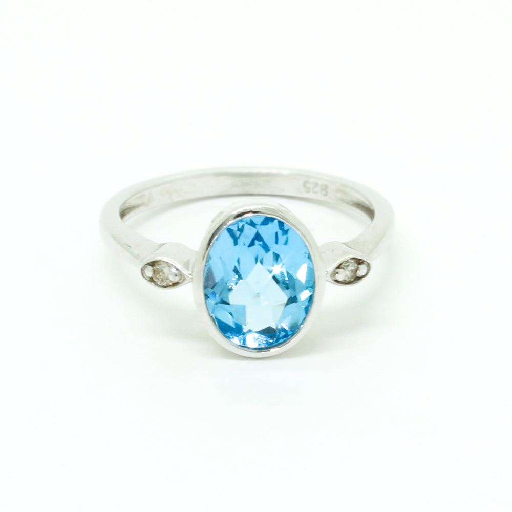 A product photo of a silver ring with a large bezel-set topaz centre stone sitting on a white background. The silver band is simple and smooth, connecting on either side of a vertically-oriented oval-cut topaz stone surrounded by a solid frame of silver. Before the band reaches the centre stone, it splits into two small horizontal silver oval frames on each side, each housnig a small diamond. The topaz jewel is almost the colour of a tropical sea, reflecting light across its multi-faceted surface.