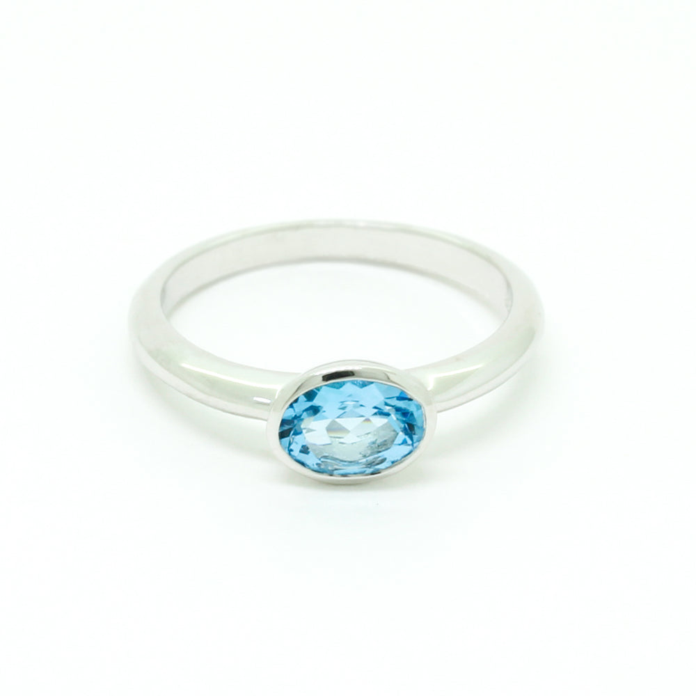 
            
                Load image into Gallery viewer, A product photo of a silver ring with a bezel-set topaz centre stone sitting on a white background. The silver band is simple and smooth, connecting on either side of a horizontally-oriented oval-cut topaz stone surrounded by a solid frame of silver. The topaz jewel is almost the colour of a tropical sea, reflecting bright blue light across its multi-faceted surface.
            
        