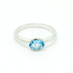 A product photo of a silver ring with a bezel-set topaz centre stone sitting on a white background. The silver band is simple and smooth, connecting on either side of a horizontally-oriented oval-cut topaz stone surrounded by a solid frame of silver. The topaz jewel is almost the colour of a tropical sea, reflecting bright blue light across its multi-faceted surface.