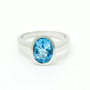 A product photo of a silver ring with a large bezel-set topaz centre stone sitting on a white background. The silver band is simple and smooth, connecting on either side of a vertically-oriented oval-cut topaz stone surrounded by a solid frame of silver. The topaz jewel is almost the colour of a tropical sea, reflecting bright blue light across its multi-faceted surface.