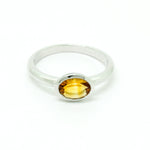 A product photo of a silver ring with a bezel-set citrine centre stone sitting on a white background. The silver band is simple and smooth, connecting on either side of a horizontally-oriented oval-cut citrine stone surrounded by a solid frame of silver. The citrine jewel is almost honey-coloured, reflecting a warm orangey yellow colour across its multi-faceted surface.
