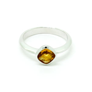 A product photo of a silver ring with a bezel-set citrine centre stone sitting on a white background. The silver band is simple and smooth, connecting on either side of a diagonally-oriented square cushion-cut citrine stone surrounded by a solid frame of silver. The citrine jewel is almost honey-coloured, reflecting a warm orangey yellow colour across its multi-faceted surface.