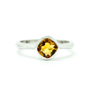 A product photo of a silver ring with a bezel-set citrine centre stone sitting on a white background. The silver band is simple and smooth, connecting on either side of a diagonally-oriented square cushion-cut citrine stone surrounded by a solid frame of silver. The citrine jewel is almost honey-coloured, reflecting a warm orangey yellow colour across its multi-faceted surface.