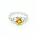 A product photo of an ornate silver ring with a circle-cut citrine centre stone sitting on a white background. The back of the silver band is simple and smooth, before splitting into four serpentine tendrils on either side that delicately coil around one another before meeting on either side of the circle-cut centre stone, which is held in place by four small silver claws. The citrine jewel is almost honey-coloured, reflecting a bright orangey yellow colour across its multi-faceted surface.