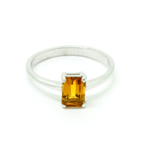 A product photo of a silver ring with an emerald-cut citrine centre stone sitting on a white background. The silver band is simple and smooth, connecting on either side of a vertically-oriented emerald-cut citrine centre stone held in placfe by four silver claws. The citrine jewel is almost honey-coloured, reflecting a warm orangey yellow colour across its multi-faceted surface.