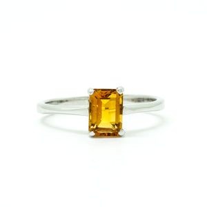 A product photo of a silver ring with an emerald-cut citrine centre stone sitting on a white background. The silver band is simple and smooth, connecting on either side of a vertically-oriented emerald-cut citrine centre stone held in placfe by four silver claws. The citrine jewel is almost honey-coloured, reflecting a warm orangey yellow colour across its multi-faceted surface.