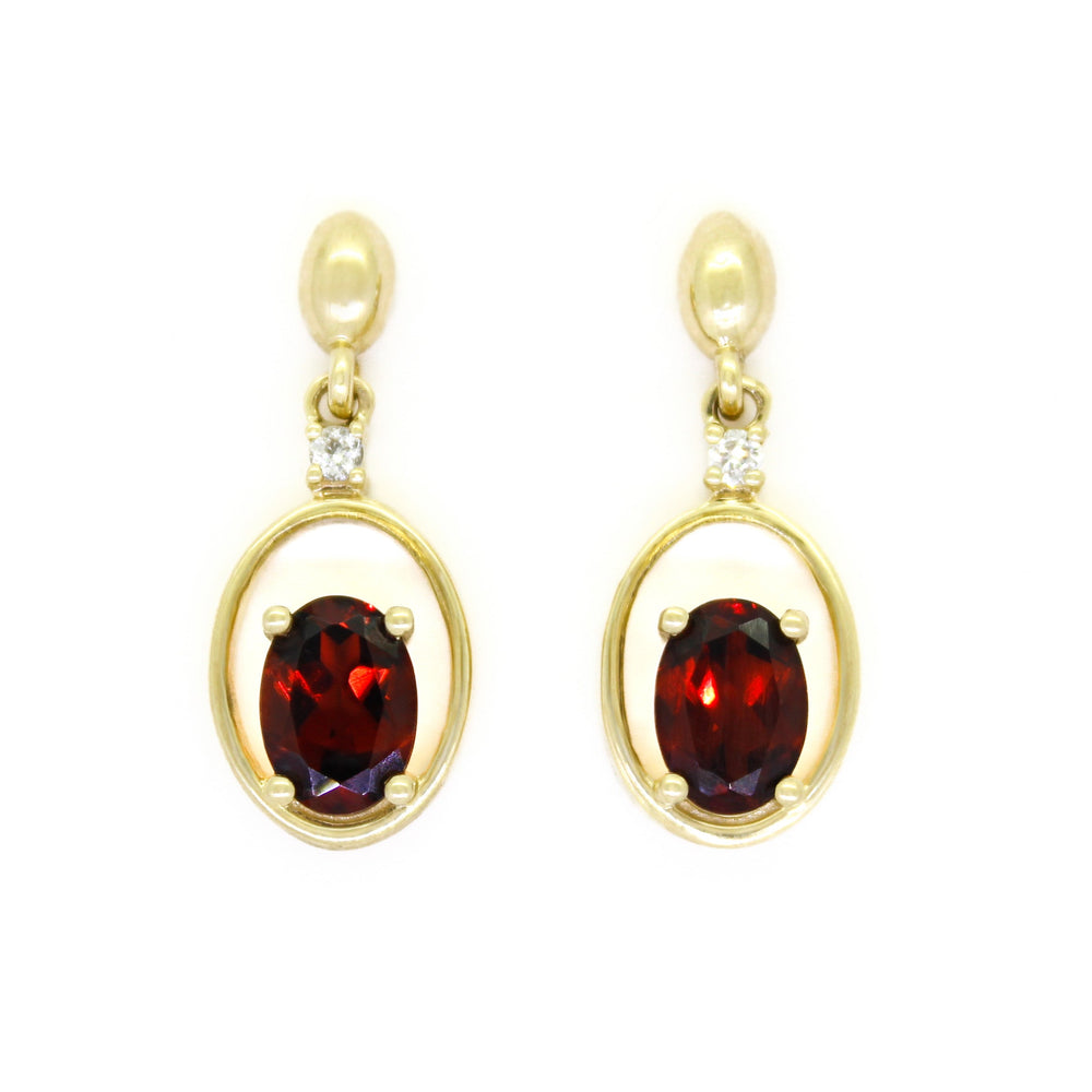 A product photo of 7x5mm Oval Garnet & Diamond Earring Studs in 9k Yellow Gold sitting on a plain white background. The stones are nestled at the bottom of golden oval hoops, with each being punctuated by a single diamond where they meet the stud. The garnets reflect sanguine hues across their multi-faceted edges.