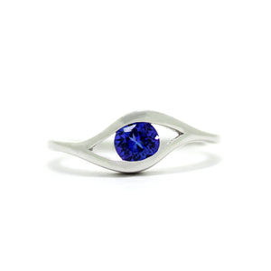 
            
                Load image into Gallery viewer, A product photo of a white gold tanzanite ring sitting on a white background. The band splits and curves around the single, round tanzanite centre stone, holding it in place like an eye. The tanzanite stone reflects violet blue and indigo colours from its many edges.
            
        