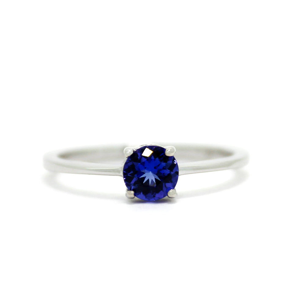 A product photo of a white gold tanzanite solitaire ring sitting on a white background. The band is smooth and simple, with the centre stone held in place by 4 small claws. The deep blue tanzanite stone reflects violet blue and indigo colours from its many edges.