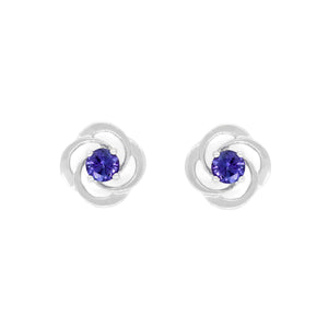 A product photo of white gold stud tanzanite earrings sitting on a white background. The small stones are framed by ornate, flowery white gold backs. The deep blue tanzanite stones reflect violet blue and indigo light from their many edges.