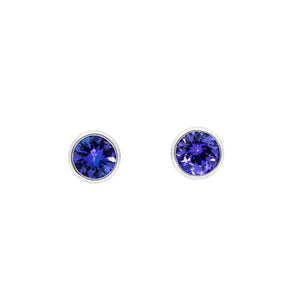 A product photo of white gold tanzanite stud earrings sitting on a white background. The bezel settings of the stones are unusually slim, drawing the viewers focus to the stones themselves. The deep blue tanzanite stones reflect violet blue and indigo light from their many edges.