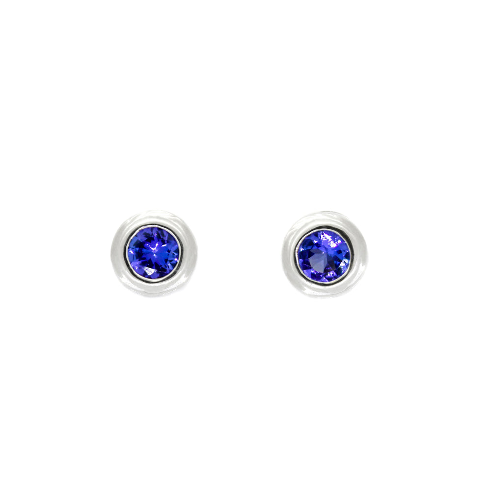 A product photo of white gold tanzanite earrings sitting on a white background. The round stones are encased in a thick layer of white gold bezel framing. The deep blue tanzanite stones reflect violet blue and indigo light from their many edges.