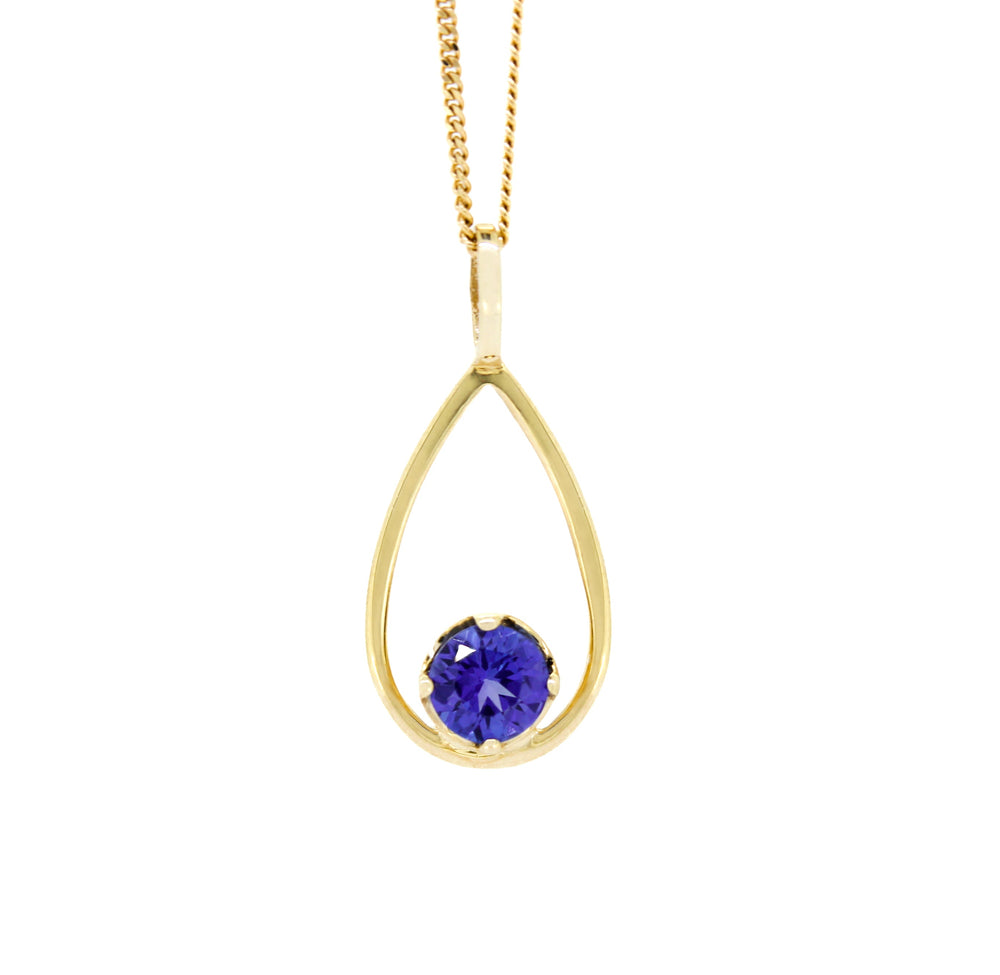A product photo of a yellow gold tanzanite pendant suspended by a chain against a white background. The deep blue tanzanite stone, held in place by 4 claws, sits at the bottom of a slim, teardrop-shaped frame of yellow gold, and reflects violet blue and indigo colours from its many edges.