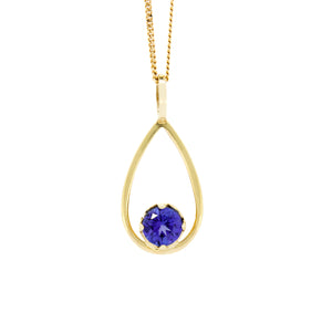 A product photo of a yellow gold tanzanite pendant suspended by a chain against a white background. The deep blue tanzanite stone, held in place by 4 claws, sits at the bottom of a slim, teardrop-shaped frame of yellow gold, and reflects violet blue and indigo colours from its many edges.