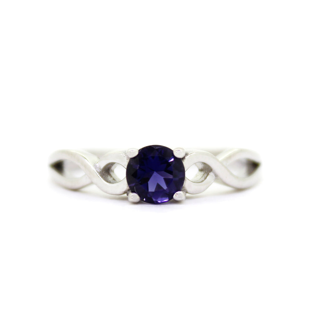 A product photo of an ornate silver ring with an iolute centre stone sitting on a white background. The silver band splits halfway along its length, becoming twisting and serpentine in appearance before meeting on either side of the deep, almost black blue 5mm stone, which is held in place by 4 silver claws.