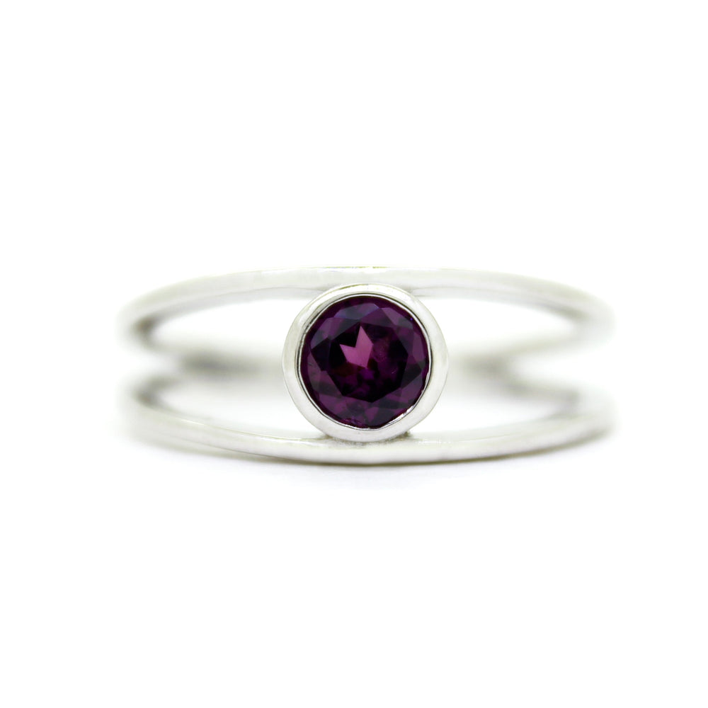 
            
                Load image into Gallery viewer, A product photo of a split-band silver ring with a bezel-set rhodalite centre stone sitting on a white background. The silver band splits at the base of the ring, separating to meet at the top and bottom of the plum-coloured round-cut centre stone, framed in a thick layer of silver, holding it in place.
            
        