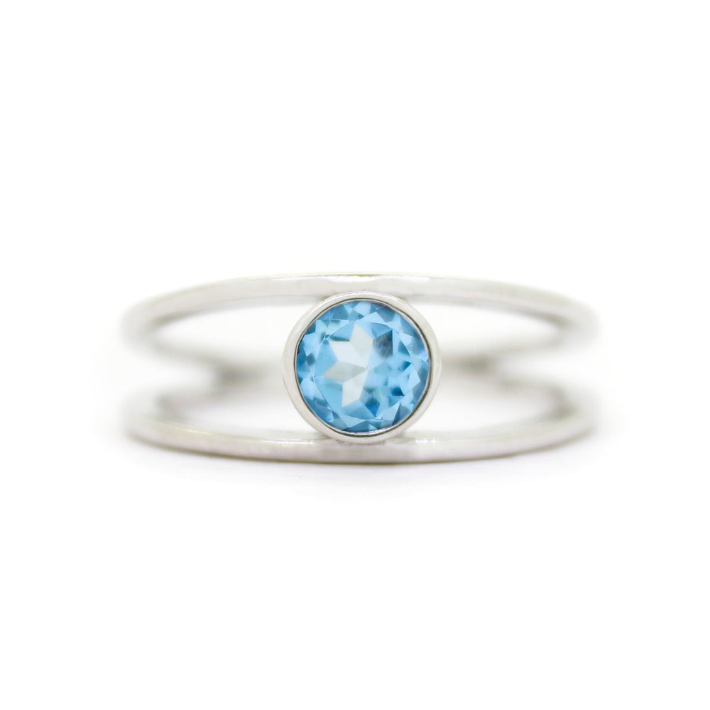 A product photo of a split-band silver ring with a bezel-set blue topaz centre stone sitting on a white background. The silver band splits at the base of the ring, separating to meet at the top and bottom of the light blue round-cut centre stone, framed in a thick layer of silver, holding it in place.