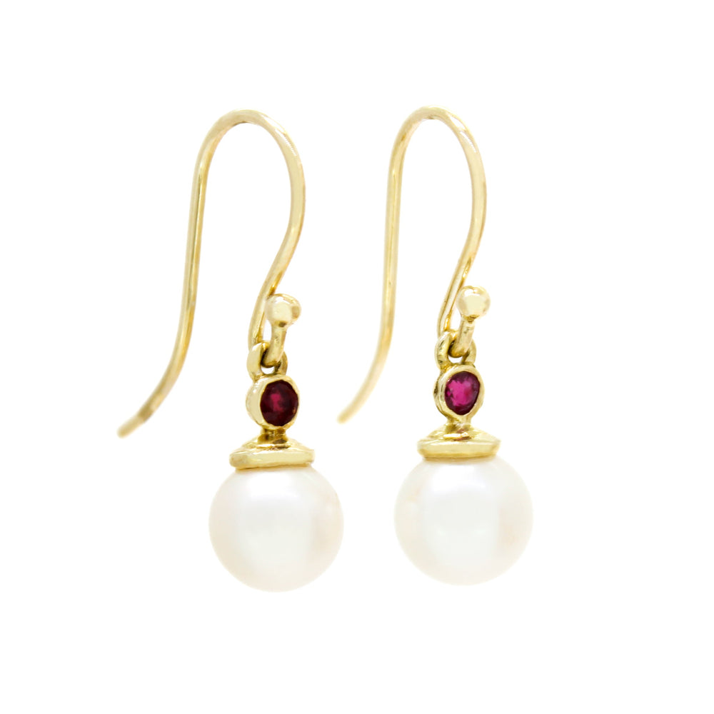 A product photo of a pair of ornate pearl drop earrings in yellow gold suspended over a white background. The pearls are white and round, with one small, pinky bezel-set ruby on each piece linking them to the long shephards hook earring.