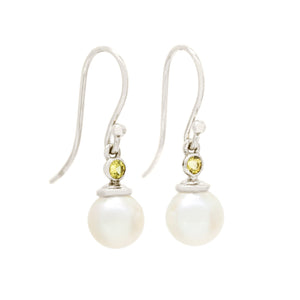 A product photo of a pair of ornate pearl drop earrings in white gold suspended over a white background. The pearls are white and round, with one small, yellow bezel-set beryl stone on each piece linking them to the long shephards hook earring.