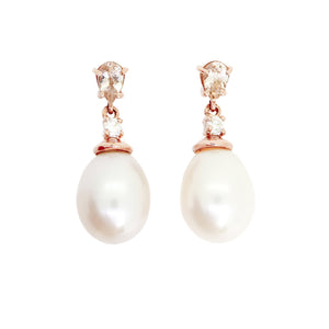 A product photo of a pair of ornate pearl drop stud earrings in rose gold suspended over a white background. The pearls are large and creamy white, with one small diamond followed by a pear-shaped pink morganite on each piece linking them to the back of the golden stud.