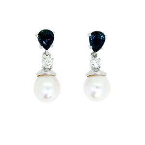 A product photo of a pair of ornate pearl drop stud earrings in white gold suspended over a white background. The pearls are creamy white and round, with one small diamond followed by a pear-shaped sapphire on each piece linking them to the back of the golden stud.